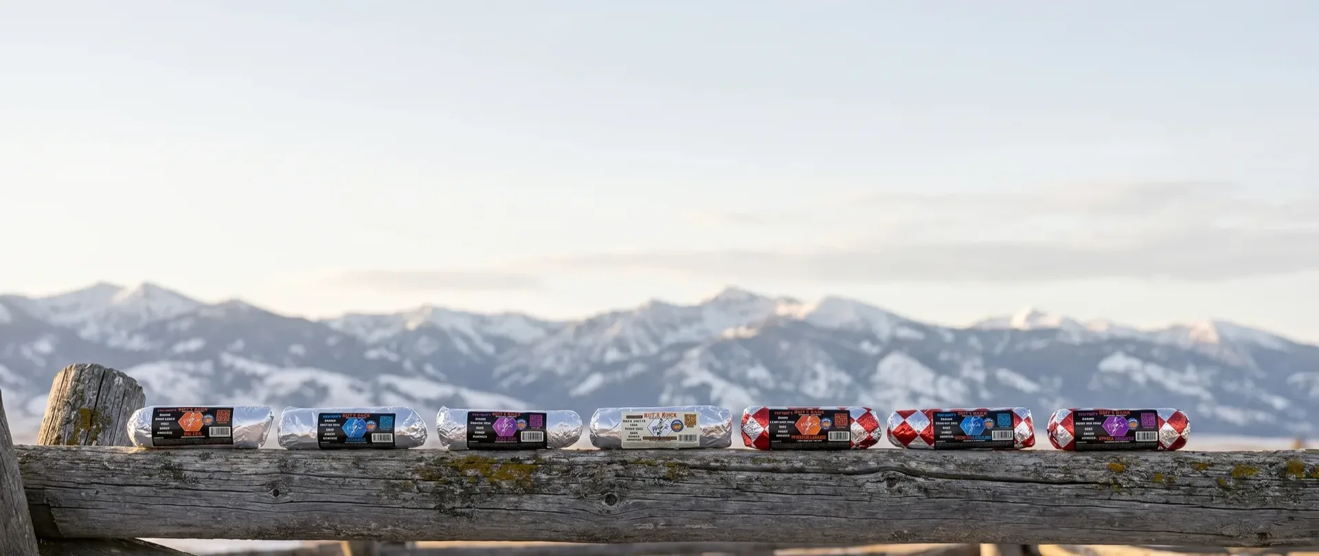 The full Rut'n Buck breakfast burrito lineup — all seven SKUs in their labeled foil wraps — resting on a weathered fence rail with the snow-dusted Bozeman mountains behind